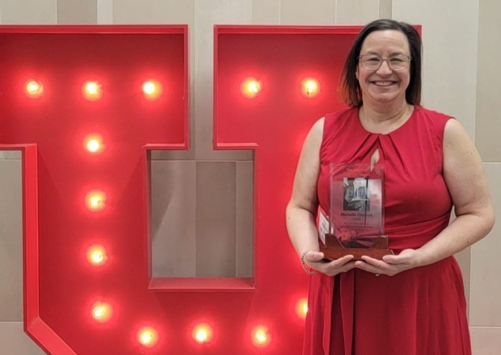 Michelle Oldroyd standing in front of a large, lit up U from the University of Utah. She is wearing a red dress and holding the Spirit of Quinney award.
